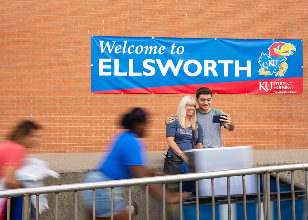 A student and their parent, who is wearing a Jayhawk shirt, take a selfie in front of a banner that reads "Welcome to Ellsworth". Students push carts in the foreground, and are blurred by motion. 