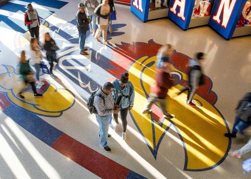 An aerial view of a parent stopping to check their phone with their student. Beneath their feet is a painted Jayhawk. Other people move around them as they pause.